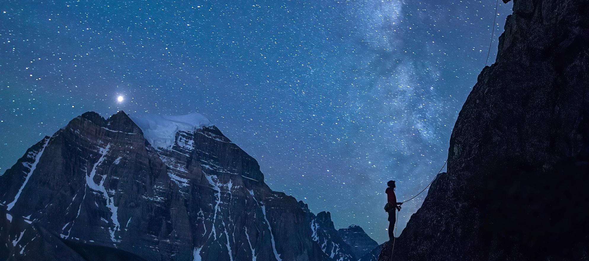 Mountaineer stands at the base of a mountain on a starry evening.
