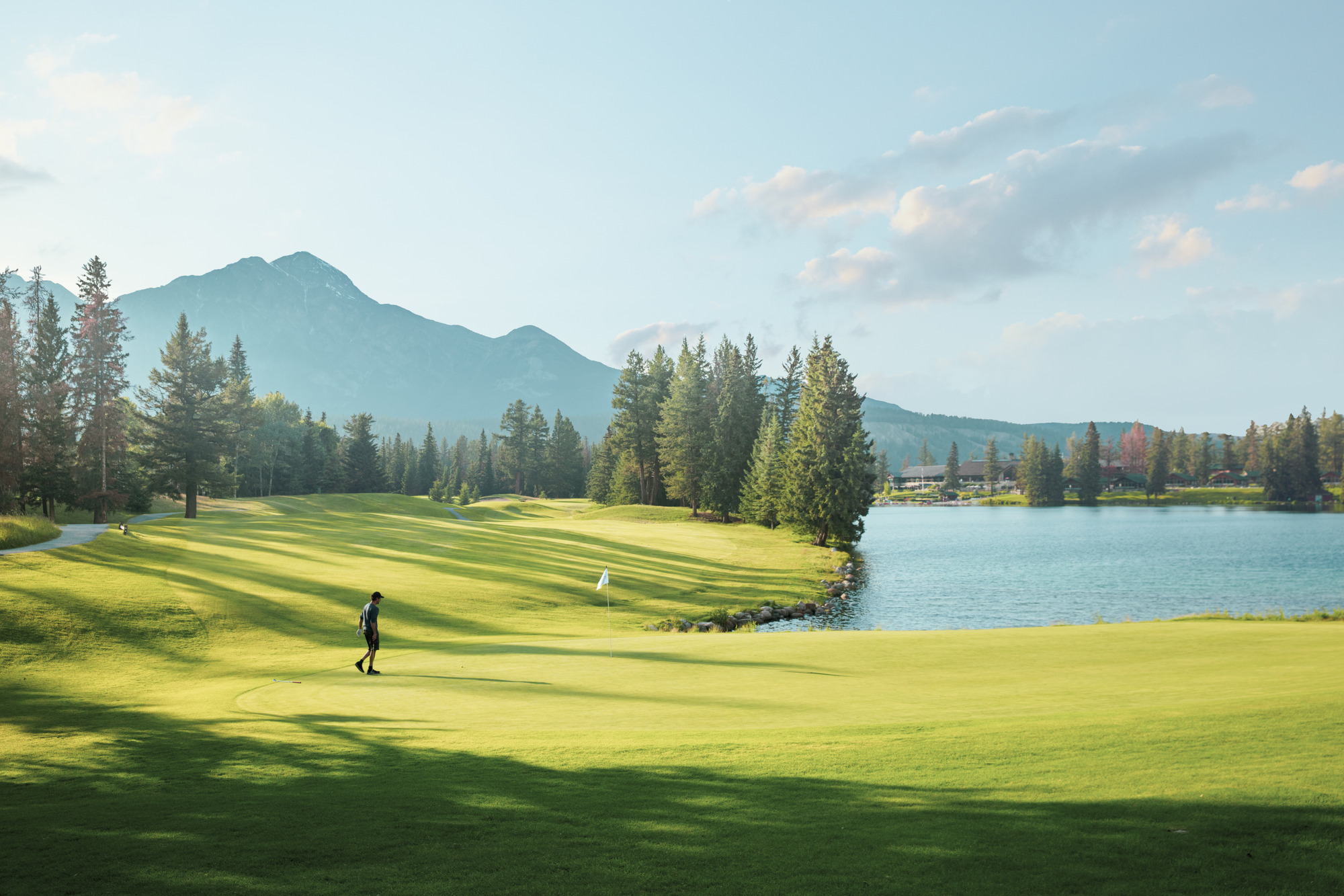 (C) Jacob Sjöman. jacob@sjomanart.com A golfer is on the green beside a lake with Jasper Park Lodge in the background.