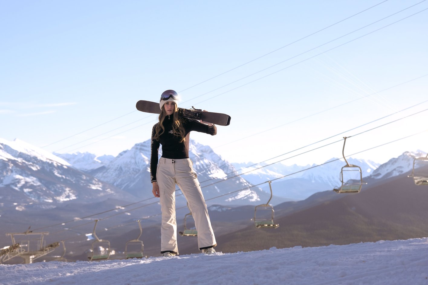 A woman stands with her snowboard on her shoulders at Marmot Basin in Jasper with the chairlift and mountains in the background